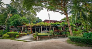 Un edificio rústico con un porche cubierto, rodeado de exuberante vegetación y un sendero de tierra que conduce a la entrada.