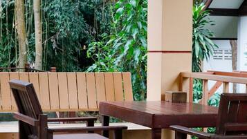 Een houten tafel en stoelen staan buiten op een betegelde vloer, omringd door groen en een houten omheining.
