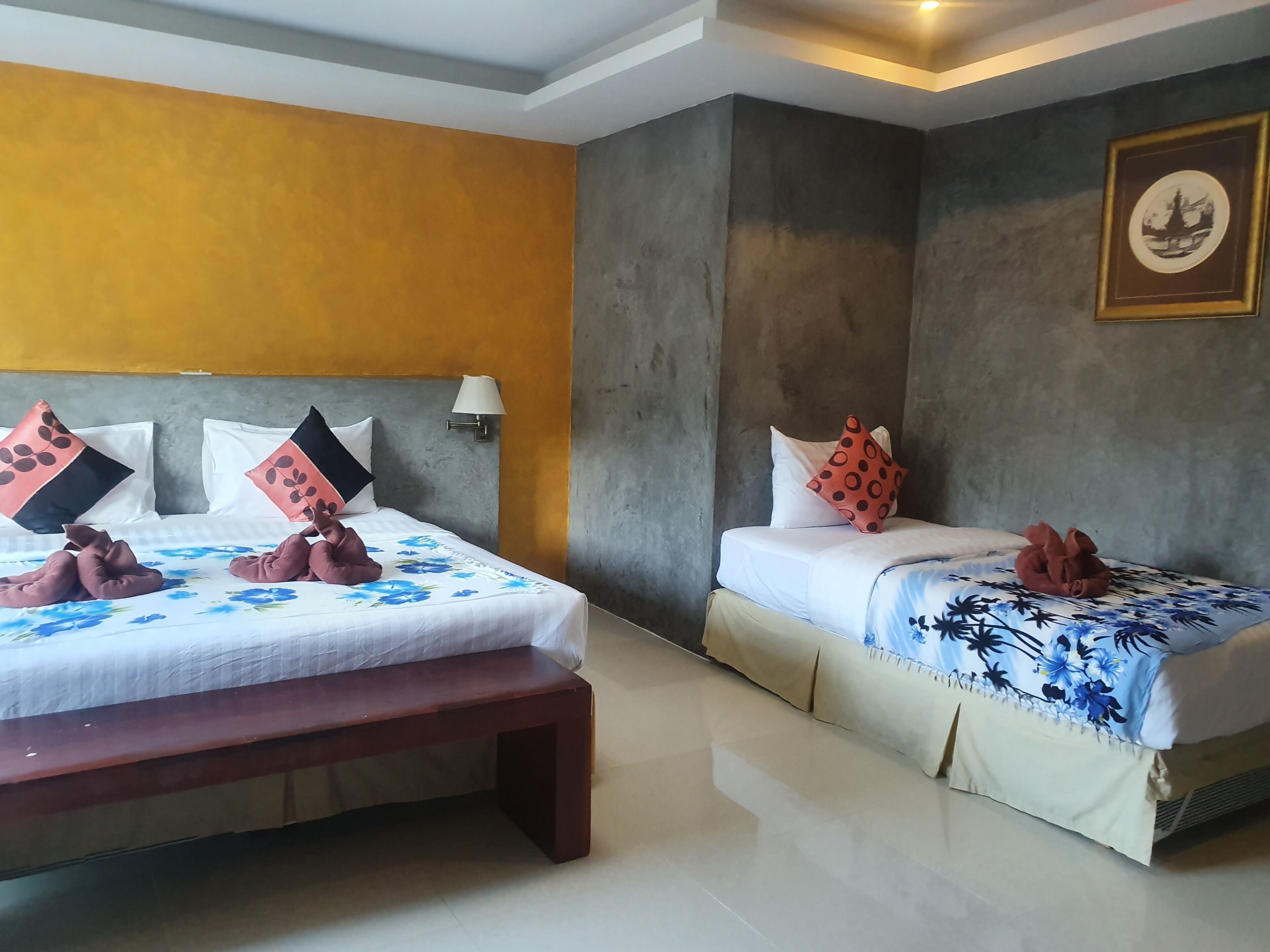 Habitación de hotel con dos camas, cada una con ropa de cama floral y almohadas decorativas. Hay un banco al pie de una cama y una lámpara de pared.