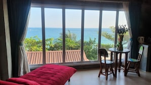 Ein Zimmer mit großem Fenster und Meerblick, einem Bett, einem Tisch mit Stühlen sowie einem kleinen Tisch mit einer Vase.