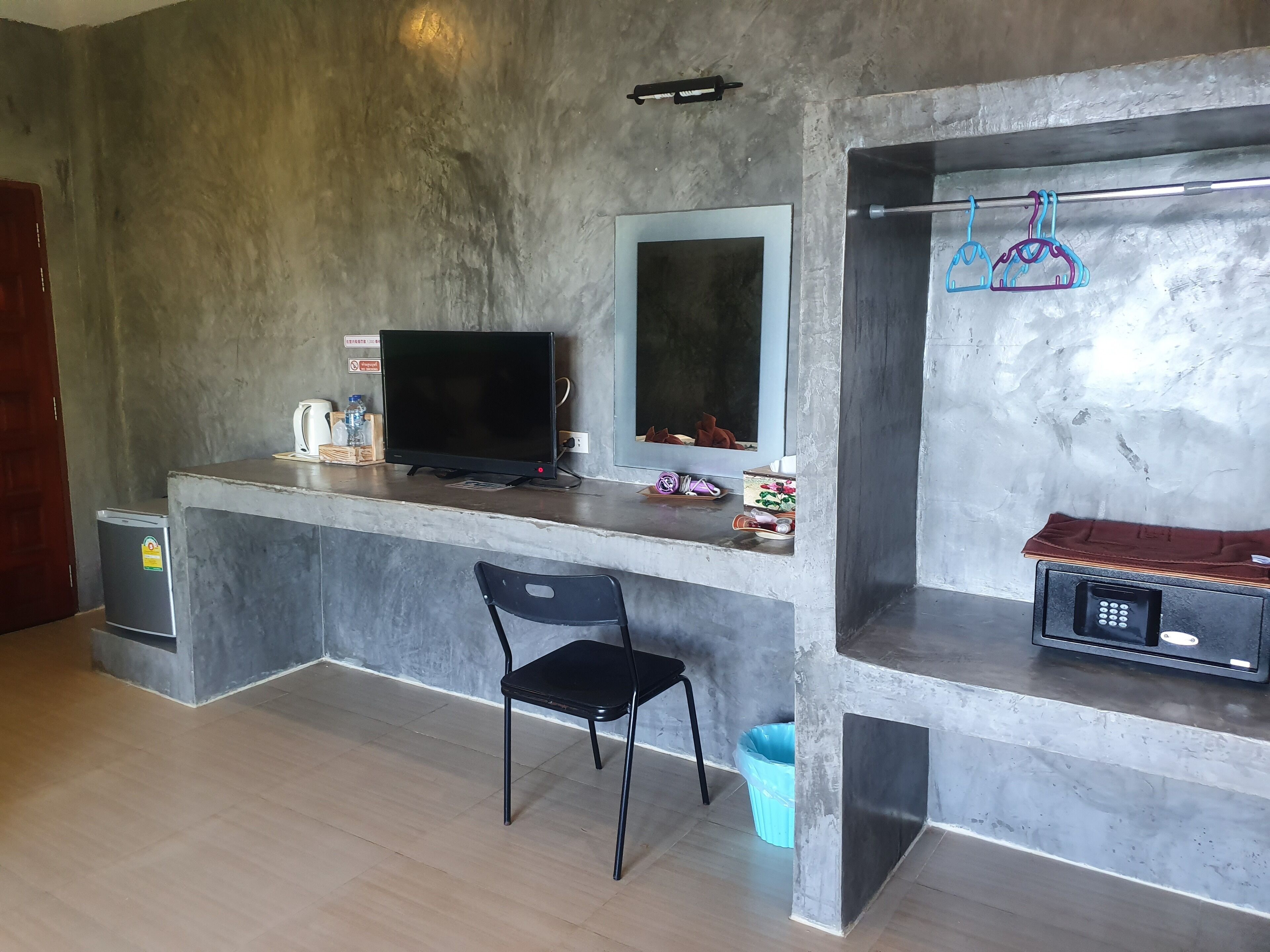A modern room with a concrete wall, a flat-screen TV, a small fridge, a desk with a chair, and a blue trash bin.