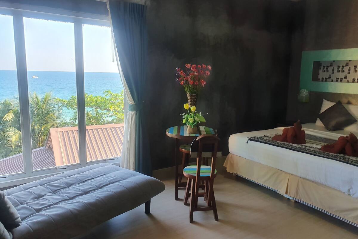 A bedroom with a bed, a chair, a small table with a vase of flowers, and a view of the ocean.