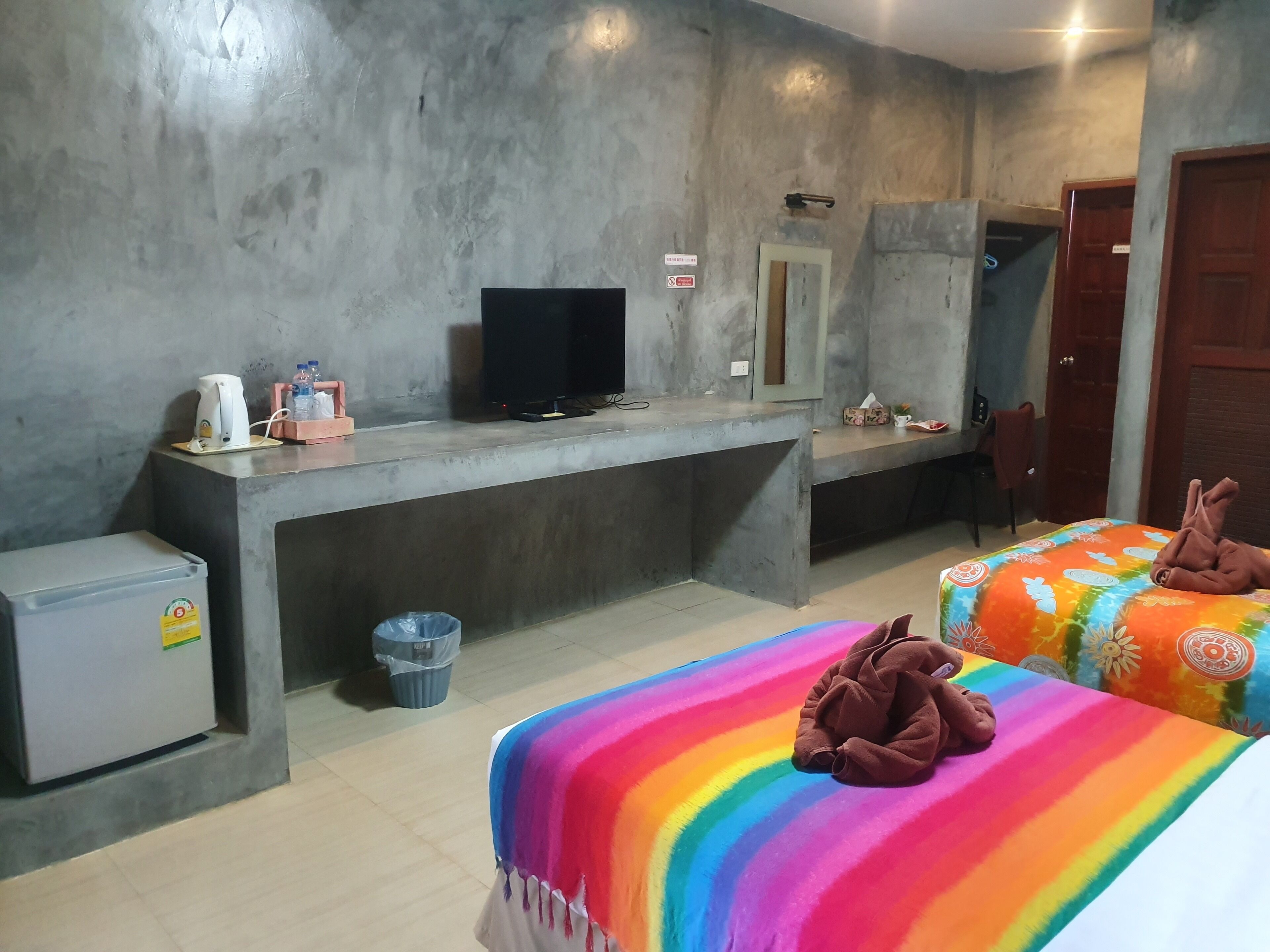 Habitación de hotel con un escritorio de hormigón, un televisor, un refrigerador pequeño, un cubo de basura y dos camas con ropa de cama colorida.