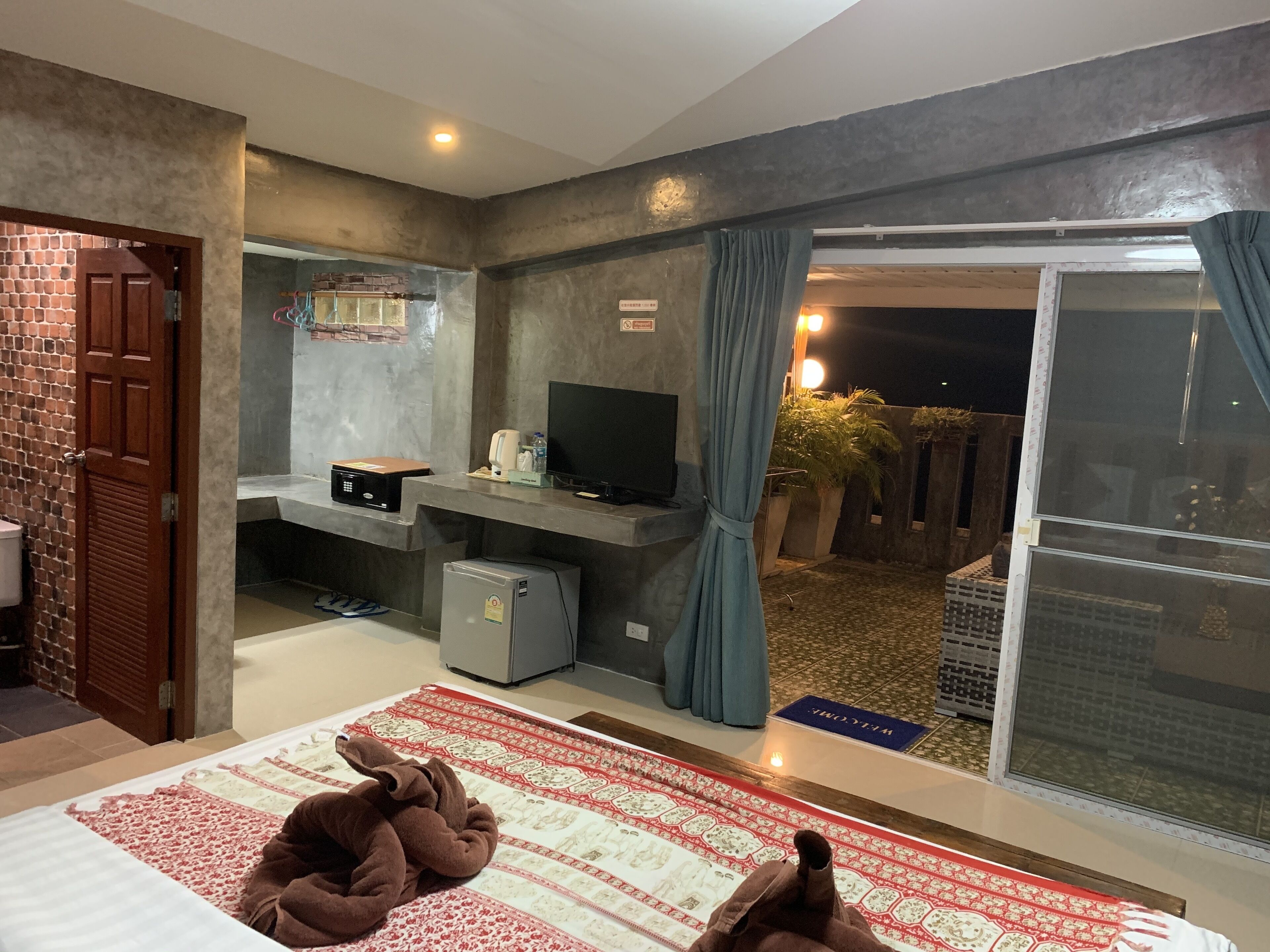 A hotel room with a bed, a television, a small refrigerator, and a sliding glass door leading to an outdoor area.