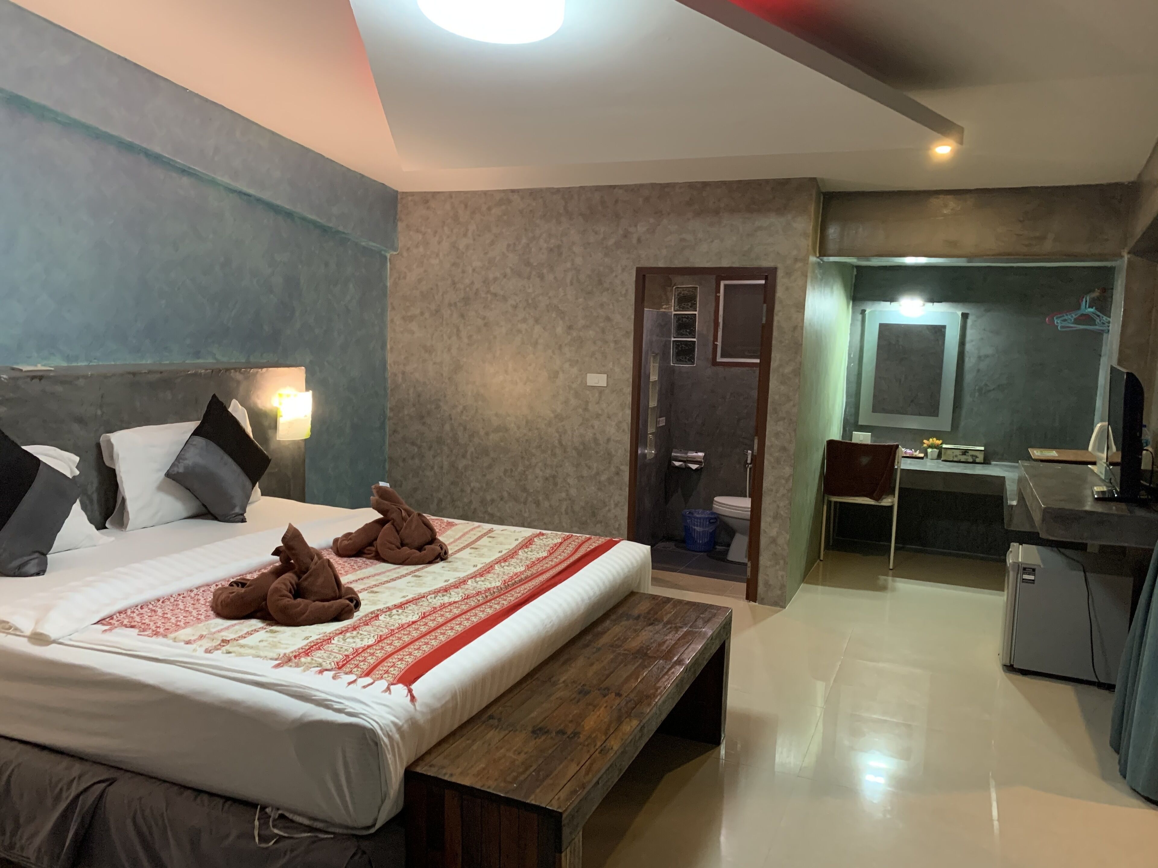A hotel room with a bed, a wooden bench, a desk, a chair, and a shower area.
