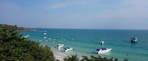 A coastal scene with multiple boats on calm waters, a sandy beach, and dense greenery in the foreground.