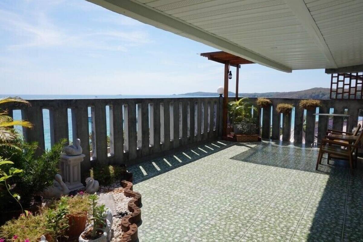 A covered patio with a view of the sea, featuring a tiled floor, potted plants, and a wooden bench.