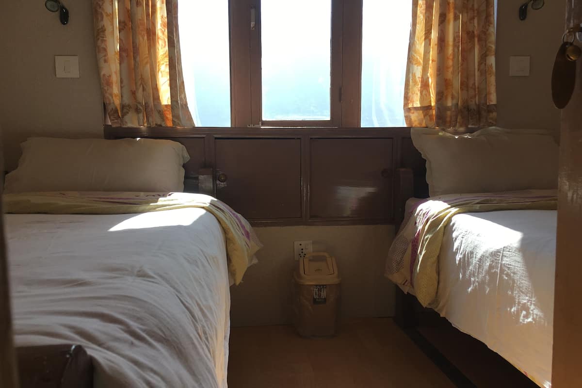 Ein Zimmer mit zwei Betten, einem fensterbelaubten Fenster, einer kleinen Mülltonne und einer Steckdose.