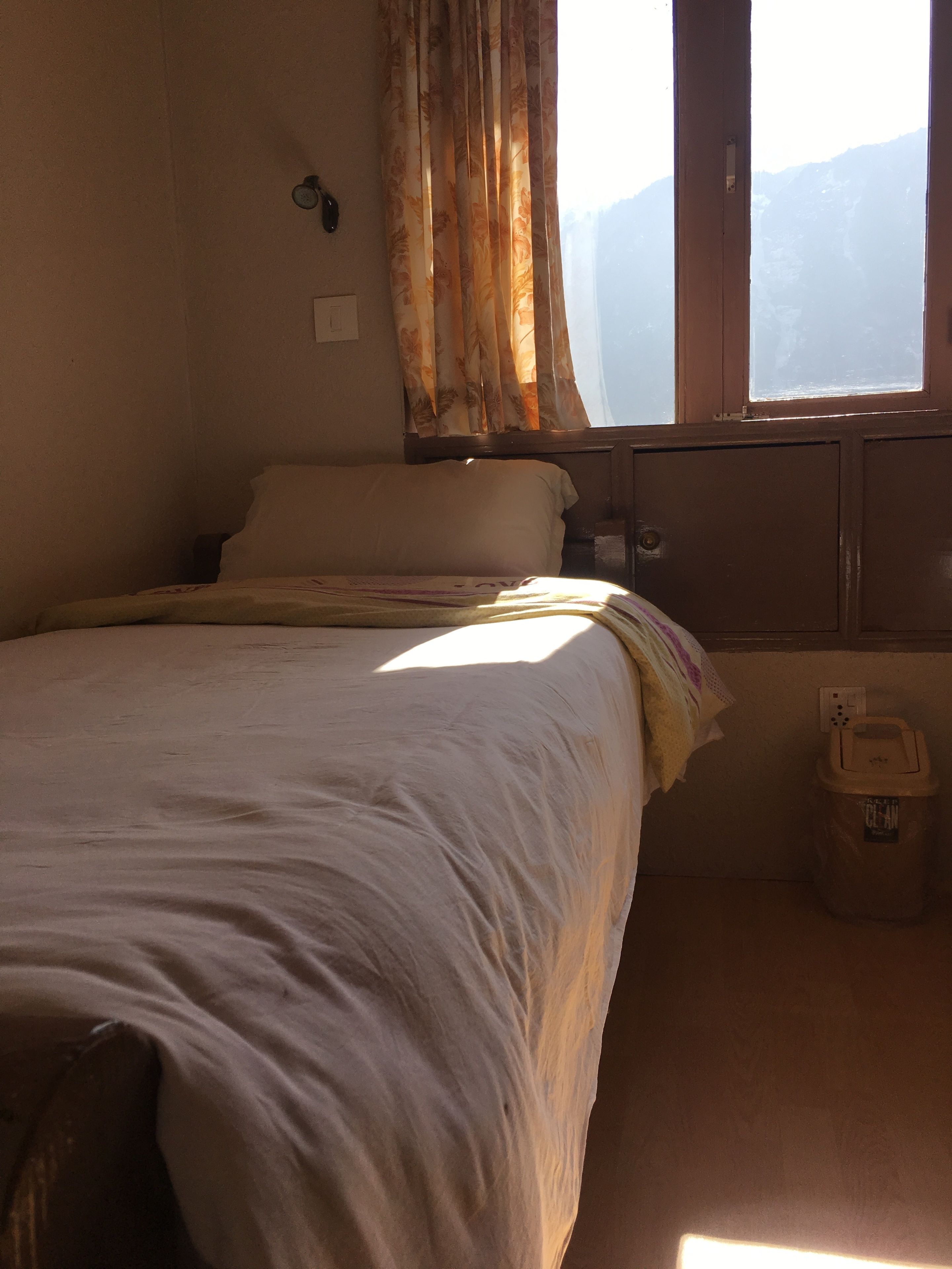 A room with a bed, a window with curtains, a wall socket, and a trash bin.