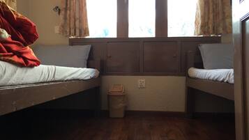 Habitación tipo dormitorio con dos literas, suelo de madera, un cubo de basura pequeño y una ventana con cortinas.