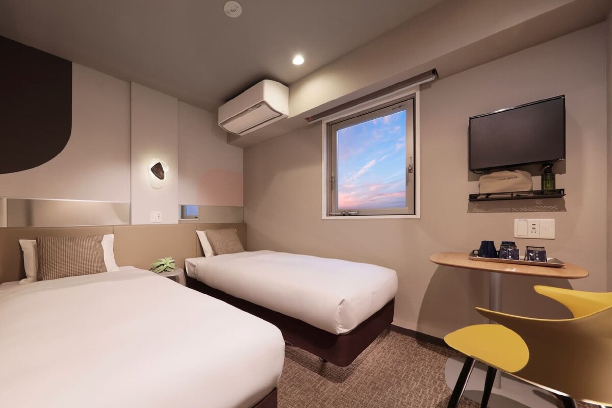 Habitación de hotel con dos camas, televisión, ventana con vistas y una mesita con tazas.