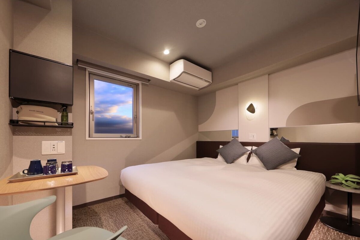 Habitación de hotel con cama, escritorio, silla, ventana y aire acondicionado.