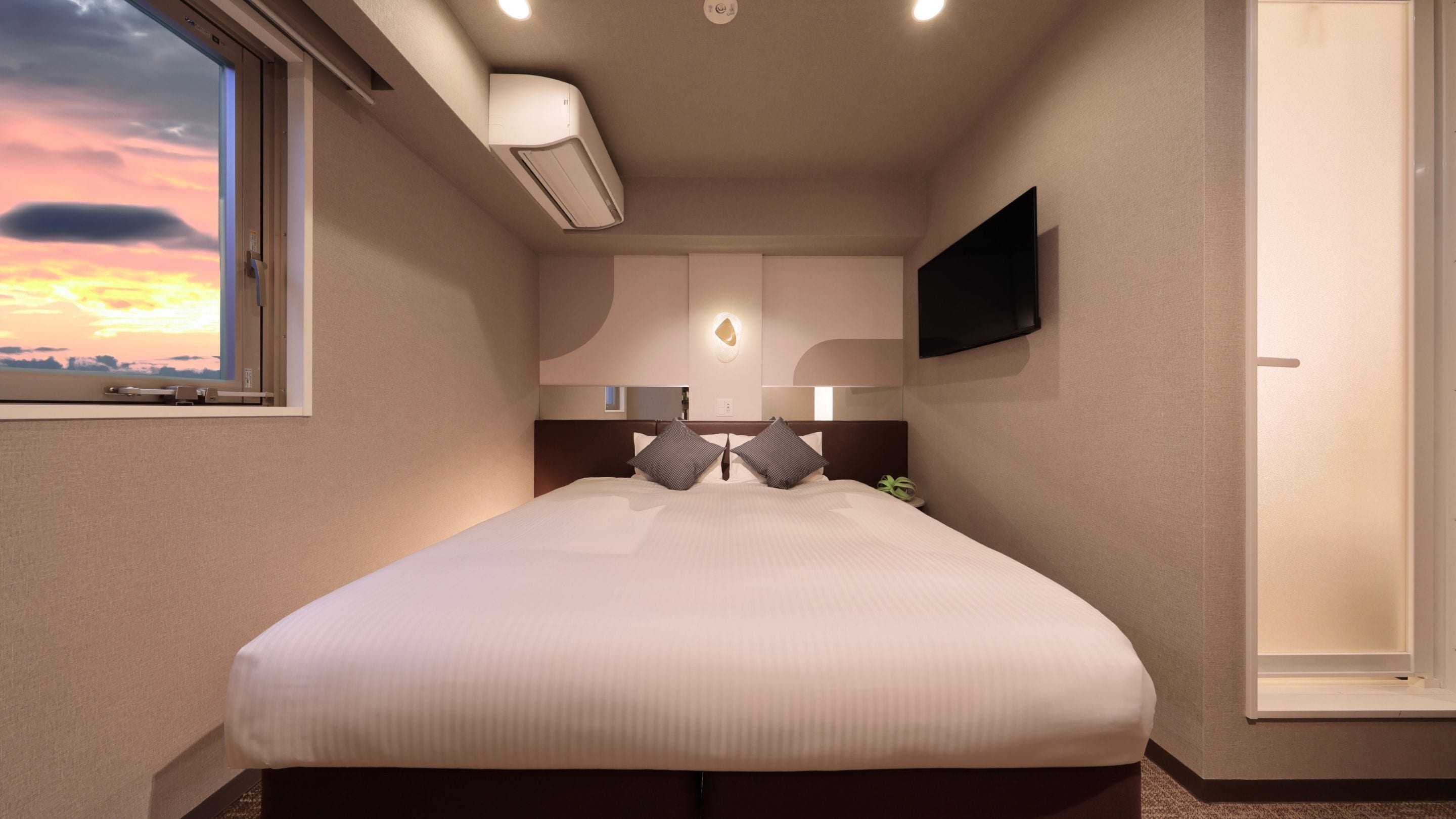 Habitación de hotel con una cama grande, un televisor de pantalla plana montado en la pared, una ventana con vista al atardecer y un equipo de aire acondicionado sobre la cama.