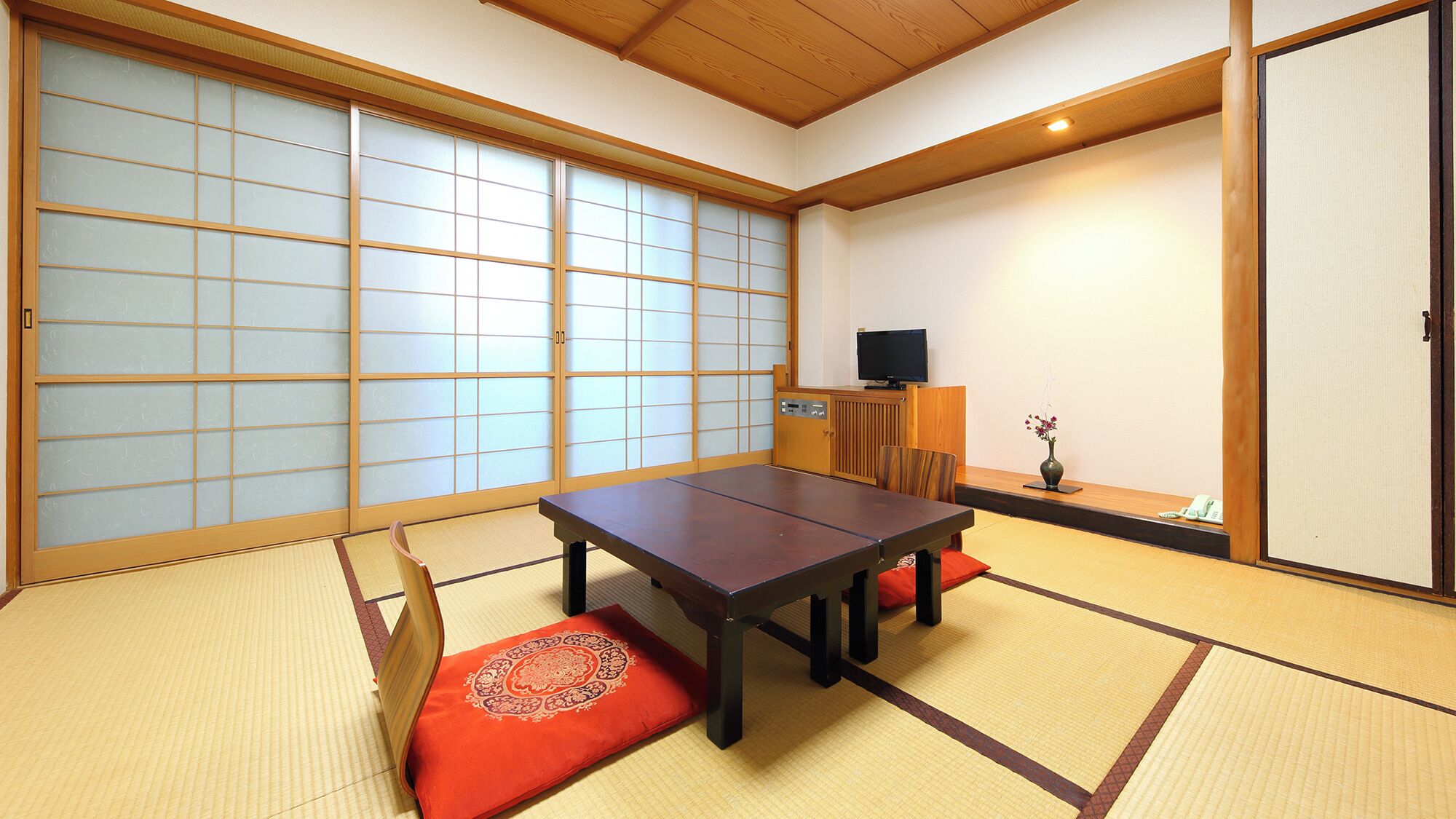 A traditional Japanese room with tatami flooring, a low table with cushions, sliding doors, and a television.