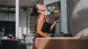 Iemand in een zwart zwempak zittend op een rieten stoel buiten, een ananas vasthoudend.