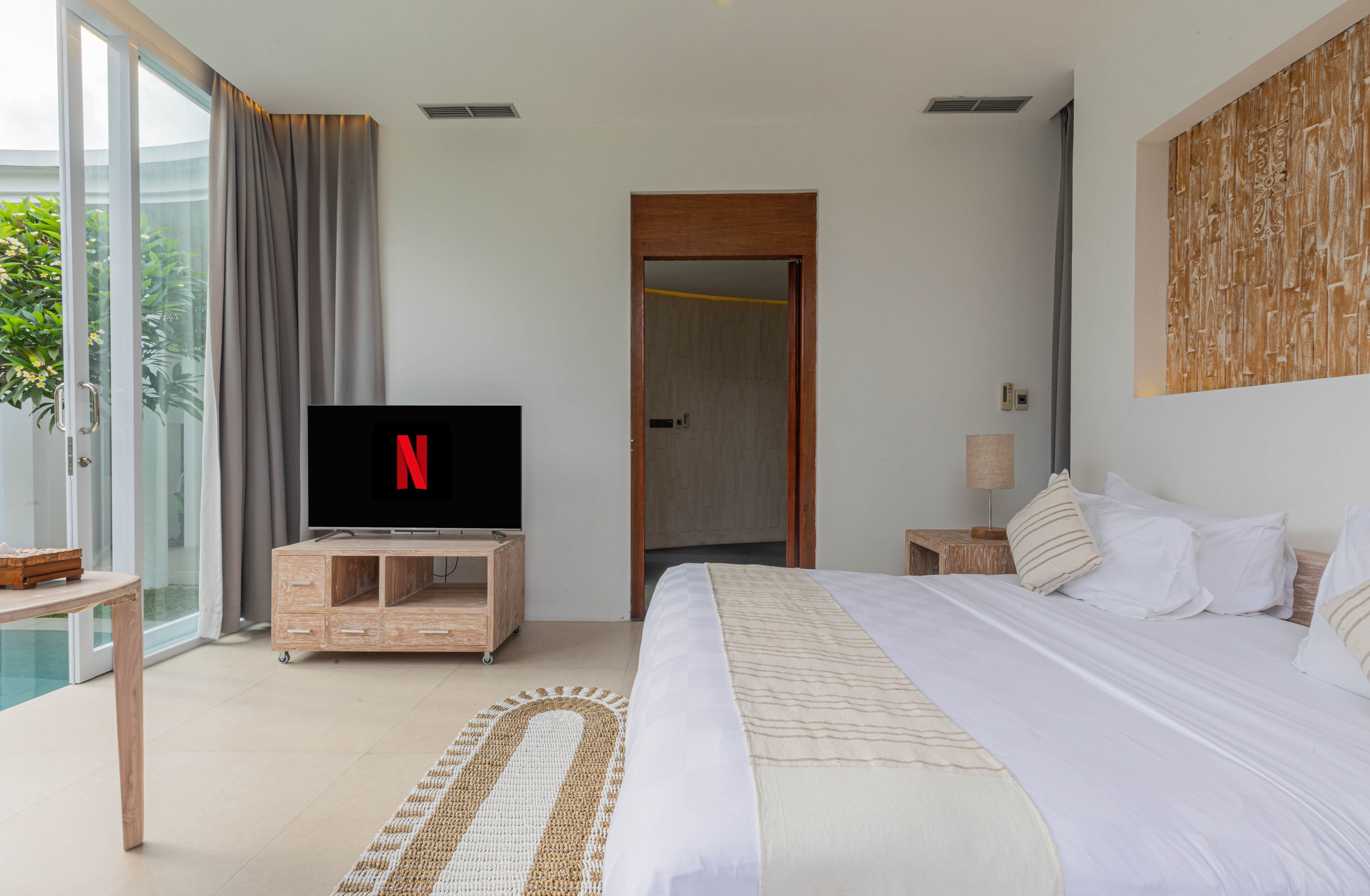 A modern hotel room with a large bed, a TV on a wooden stand, a bedside table, a lamp, and a view of the outdoors.
