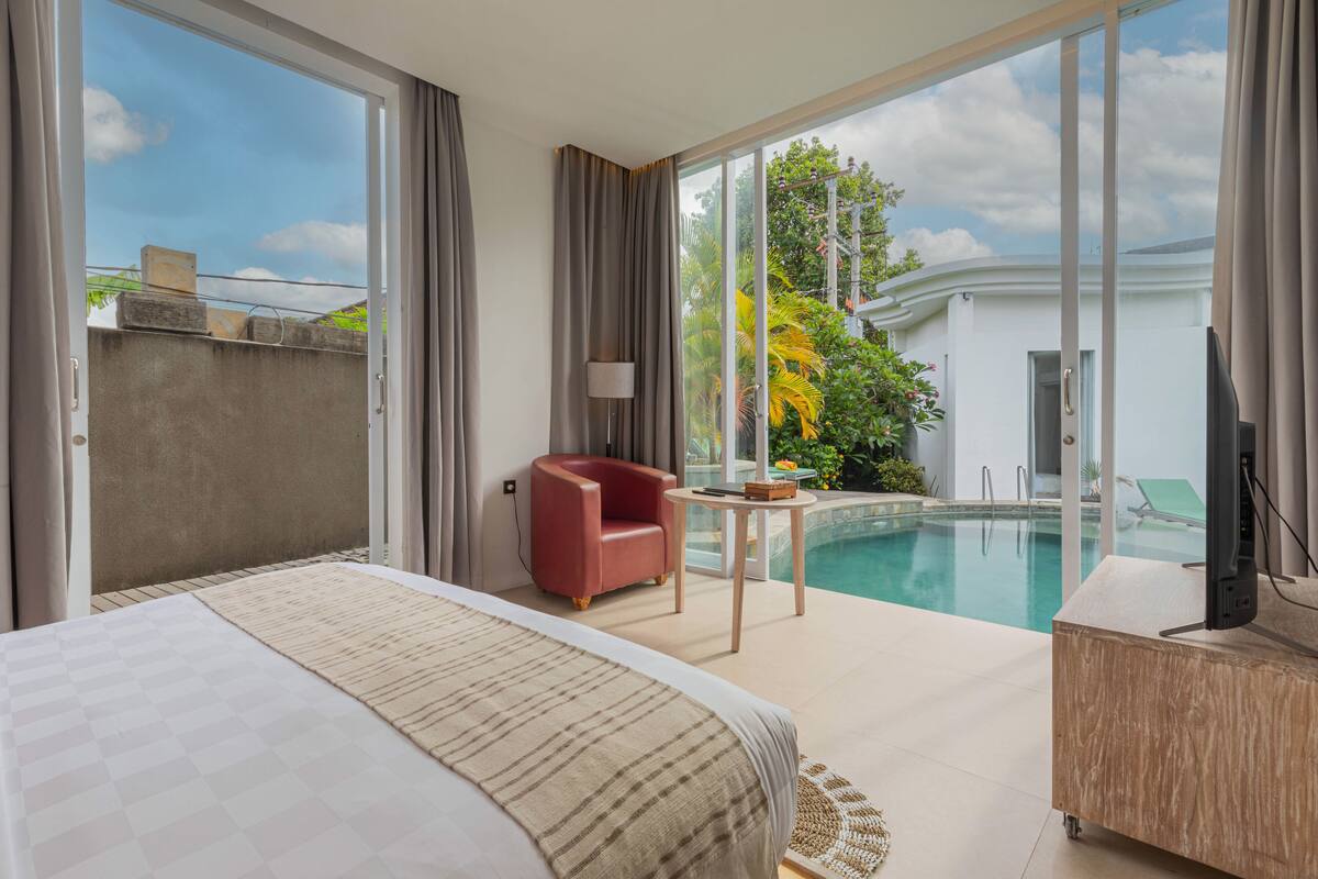 Quarto de hotel com uma cama grande, uma poltrona vermelha, uma mesa pequena, televisão e vista para a piscina.
