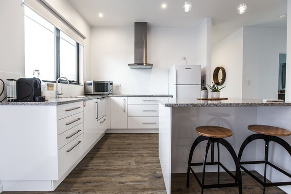 A modern kitchen with white cabinets, a stainless steel range hood, a microwave, and a refrigerator. There are two wooden stools at the island. - Island Quarters (Whitemark)
