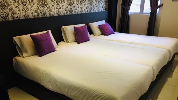 Uma cama de casal com roupa de cama branca e almofadas roxas, uma cabeceira escura e um papel de parede estampado.