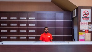Um balcão de recepção de hotel, com um homem de camisa vermelha atrás. Há vários armários marrom escuro com grelhas de ventilação, um aviso de proibido fumar e diversos avisos e documentos na parede.