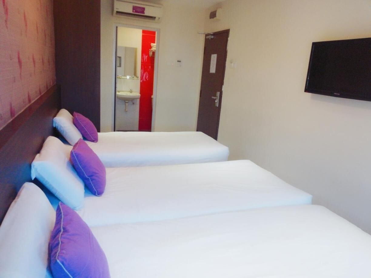 A hotel room with three beds, each with white linens and purple pillows. There is a television mounted on the wall and a door leading to a bathroom with a red accent.