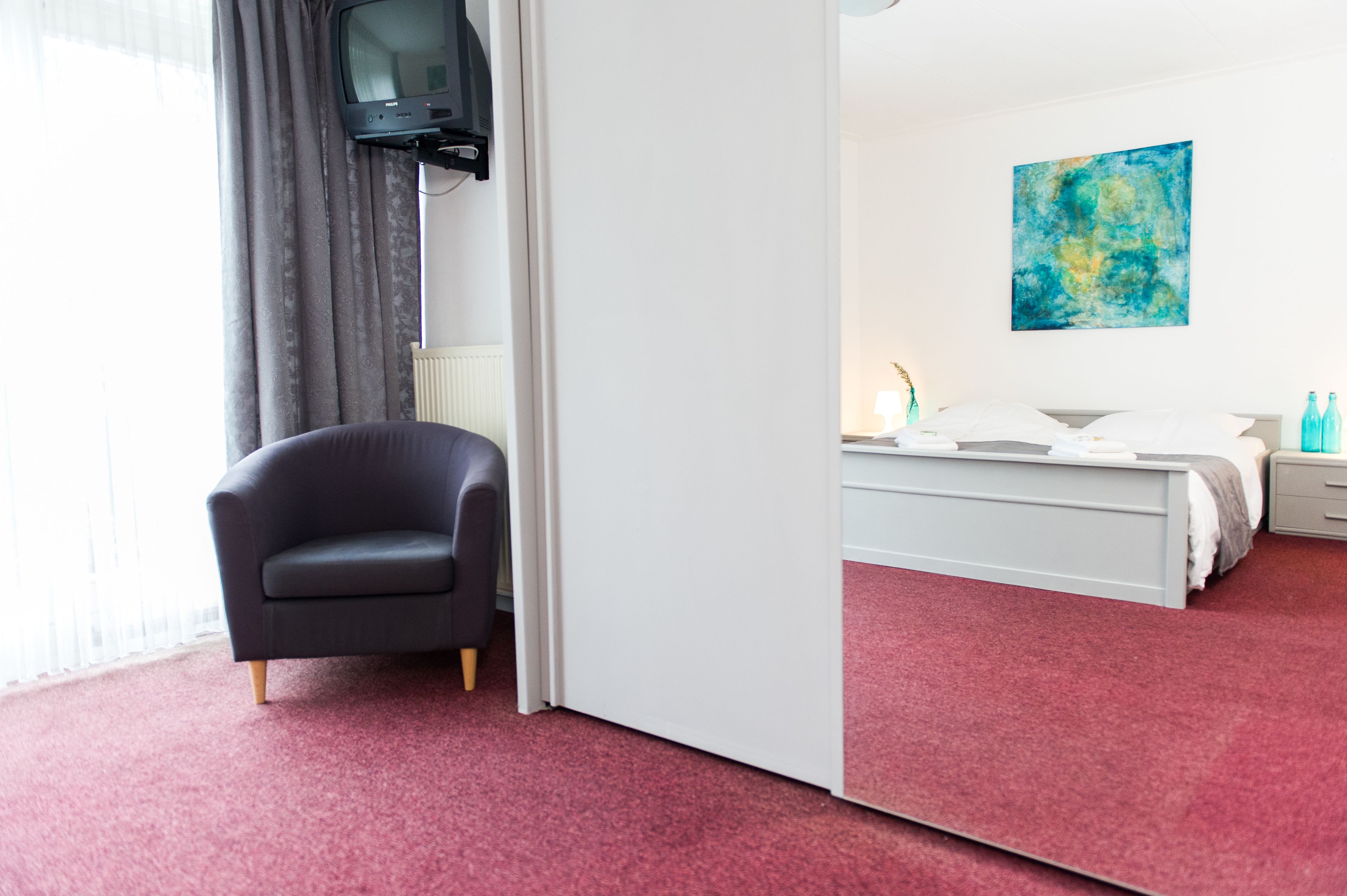 Hotelkamer met een rood tapijt, een zwarte fauteuil, een bed met wit bedlinnen, een nachtkastje, een lamp en een blauw abstract schilderij aan de muur.