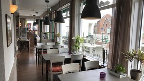 A restaurant with wooden floors, white tables and chairs, and large windows offering a view of the street. - Gasterij Berg & Dal (Slenaken)