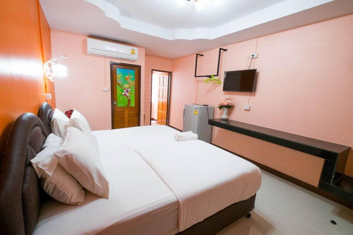 A bedroom with a bed, a TV mounted on the wall, a small desk, and an air conditioning unit.