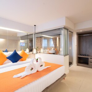A modern hotel room with a large bed, a glass partition, a bathroom with a shower, and a wooden bench.