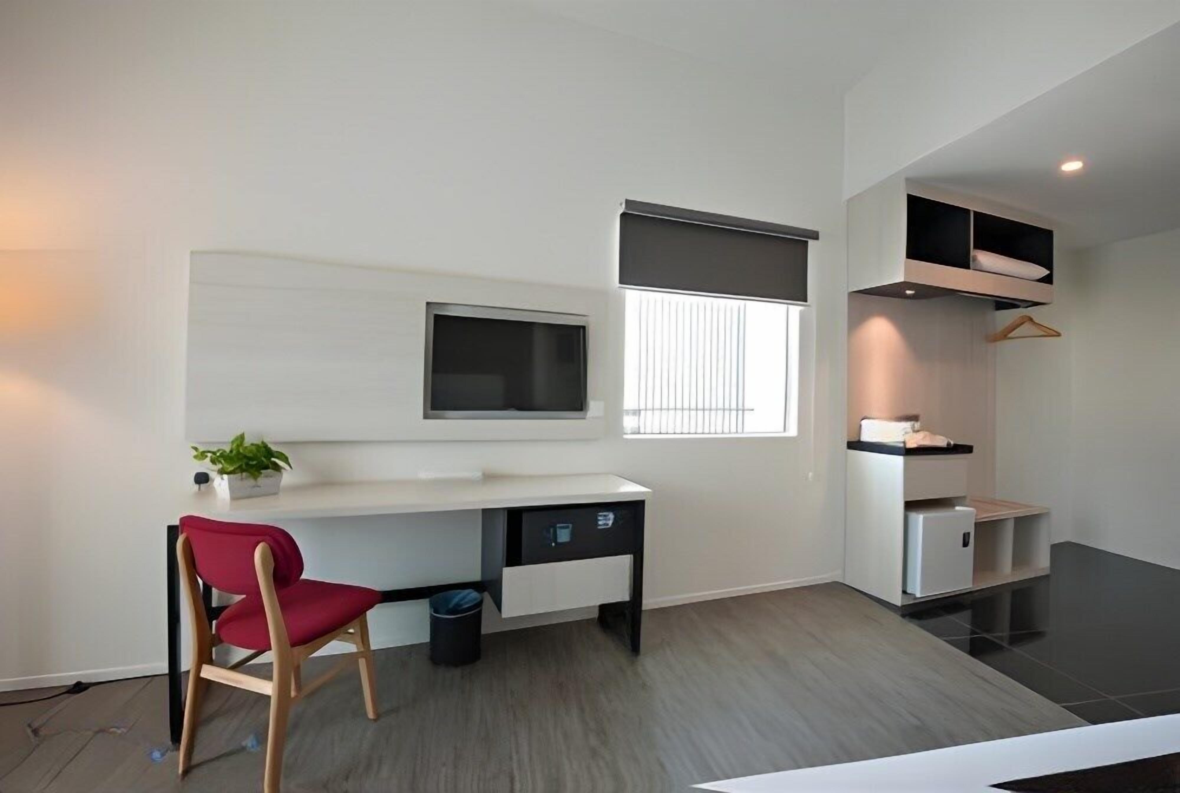 Ein modernes, minimalistisches Zimmer mit weißem Schreibtisch, einem roten Sessel, einem an der Wand befestigten Fernseher und einem Fenster mit Jalousien.