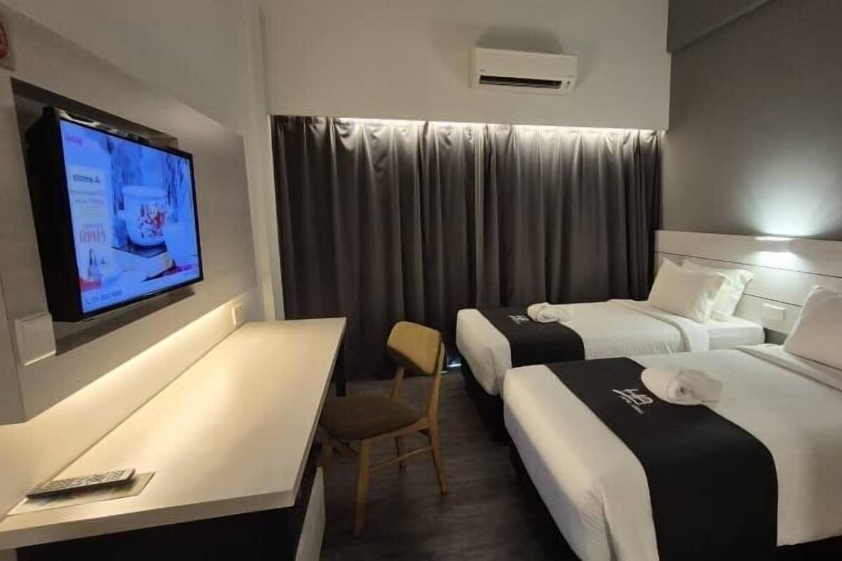 A hotel room with a TV on the left, two beds with white linens, a desk with a chair, and a dark curtain in the background.