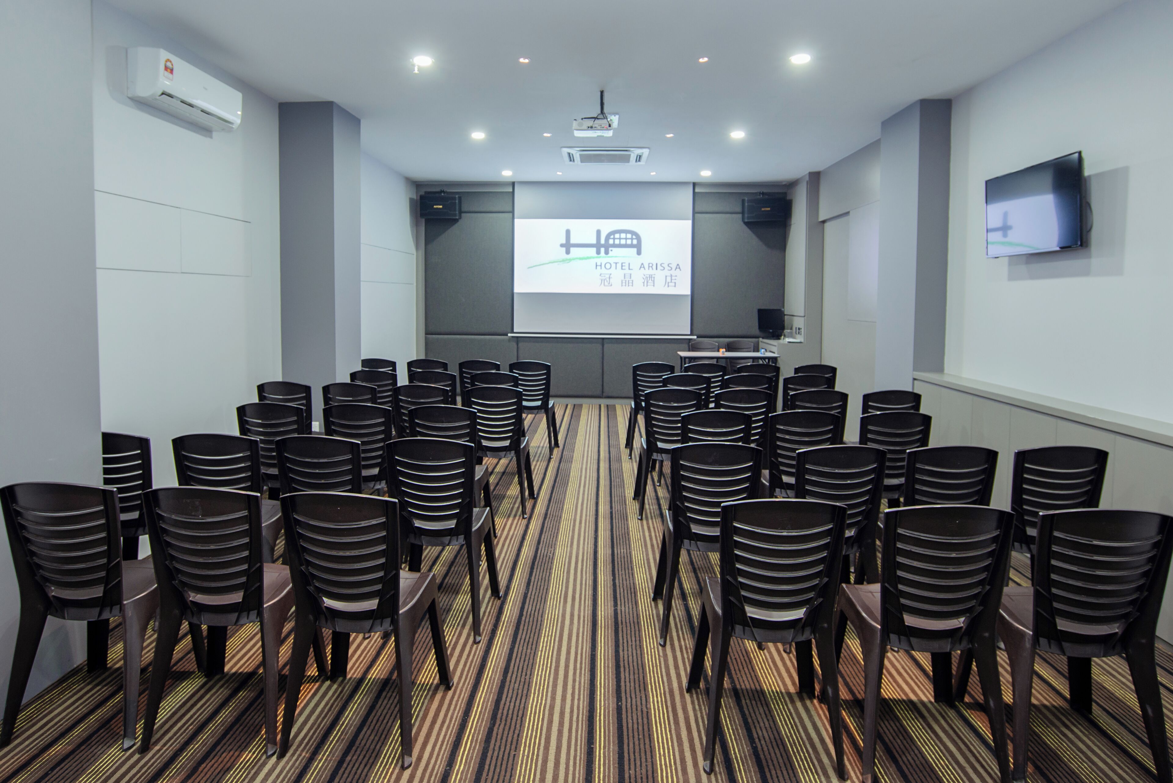Un salón de conferencias con filas de sillas, una pantalla de proyector y un televisor en la pared.