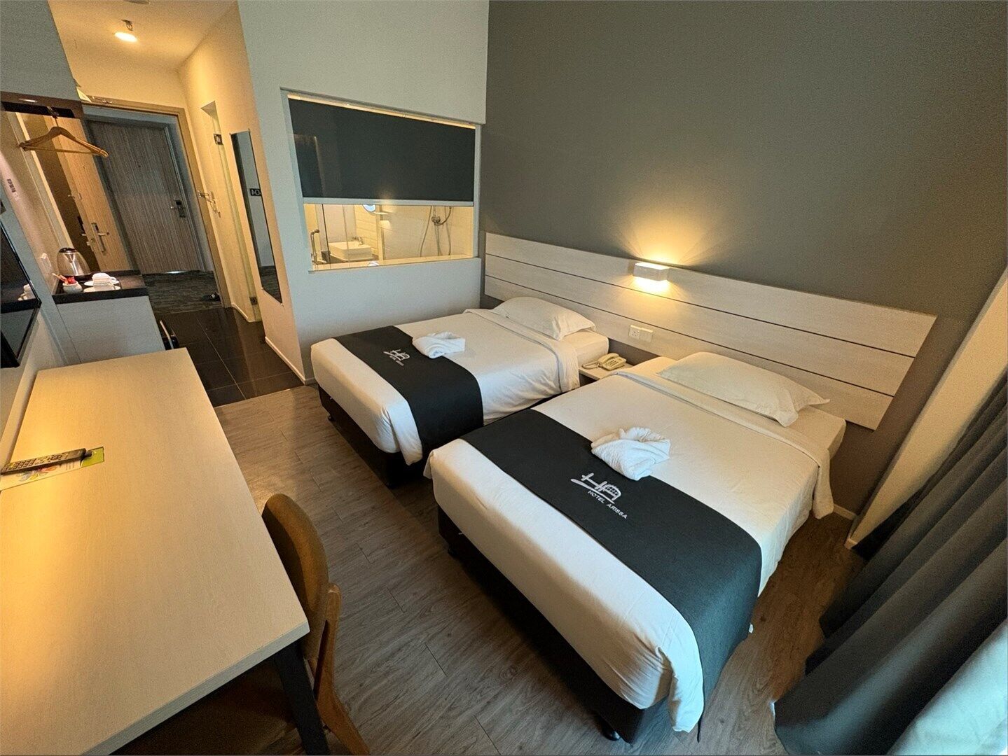 A hotel room with two beds, a desk, and a bathroom visible through a window.
