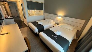 Habitación de hotel con dos camas, un escritorio y un baño visible a través de una ventana.