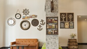 En entréhall med en brun lädersoffa, ett träbord, en marmorpelare, väggmonterade klockor, en ankardekoration och en bokhylla med böcker och inramade bilder.