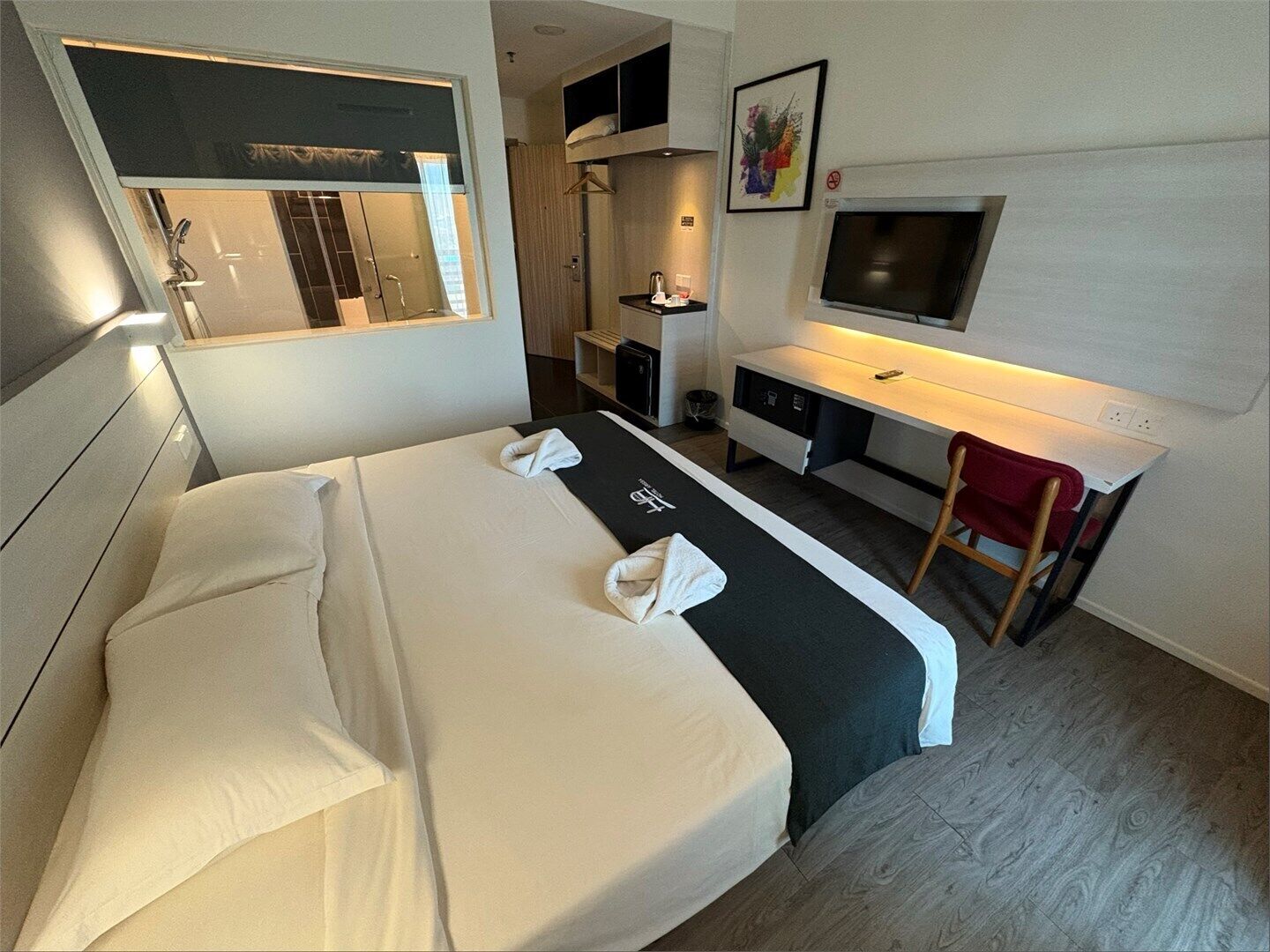 A hotel room with a bed, a desk, a chair, a TV, and a shower area.