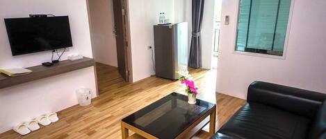 Un salón con un sofá de cuero negro, una mesa de centro de vidrio, un televisor de pantalla plana sobre una repisa de madera y un pequeño jarrón blanco con flores.