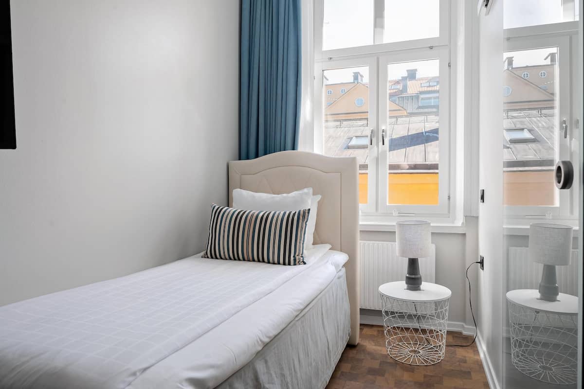 En säng för en person med en sänggavel, ett fönster med utsikt över byggnader och ett litet bord med en lampa.