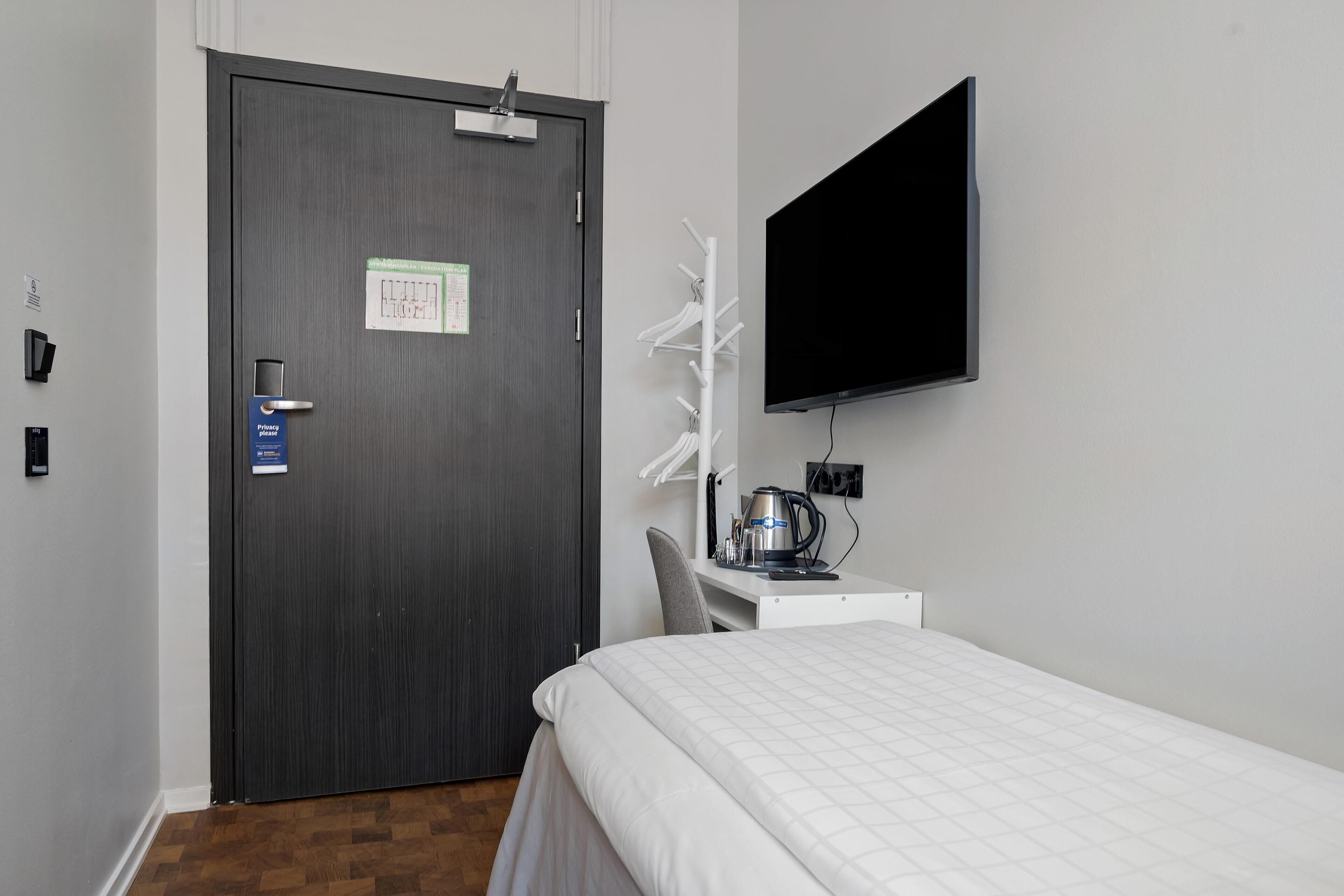 Habitación de hotel con cama, escritorio con computadora, televisión, puerta con letrero de no molestar y perchero.