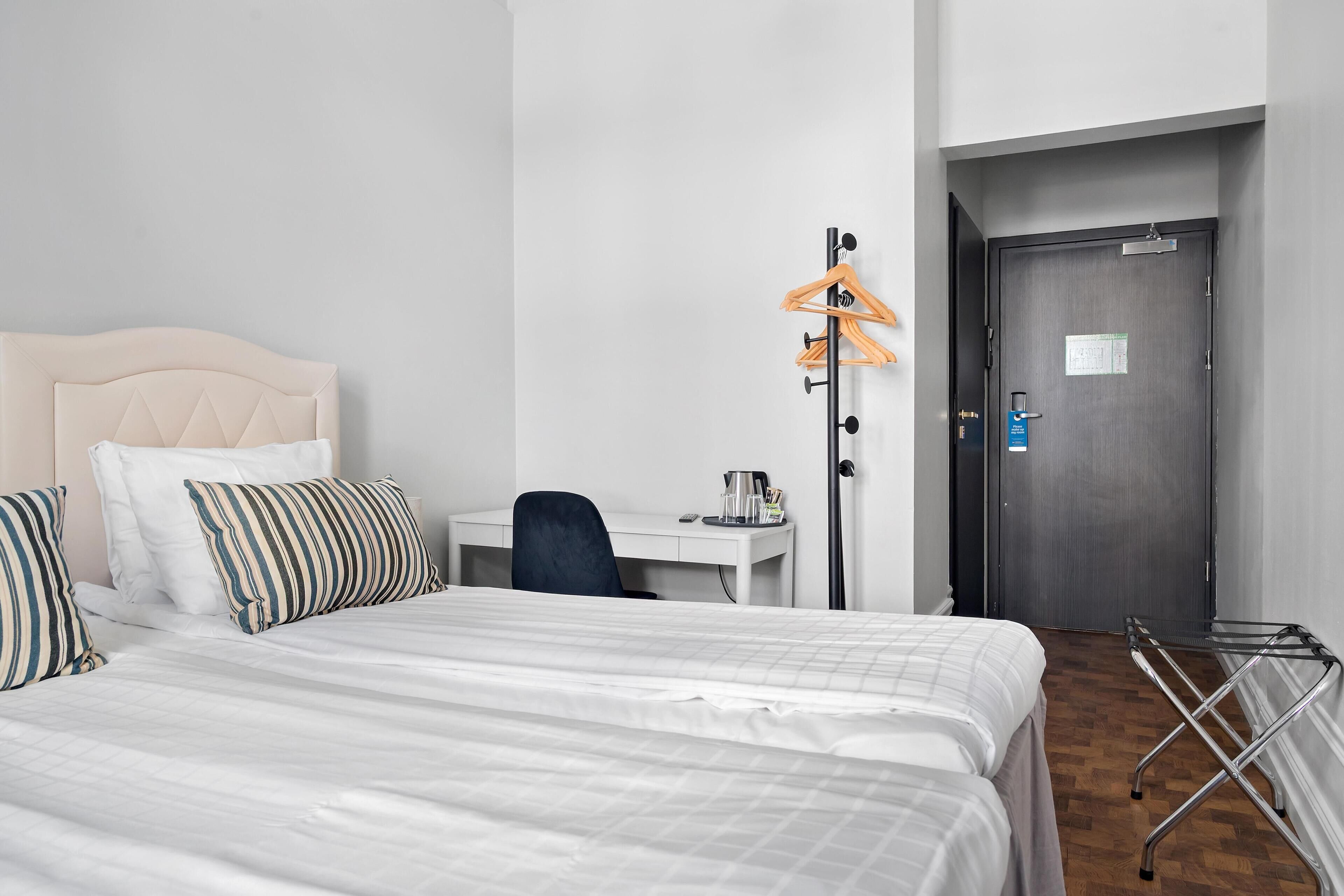 A hotel room with a bed, a desk, a chair, a coat rack, and a door.