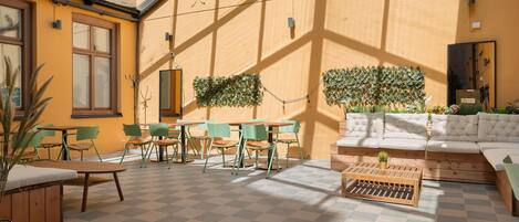Un espacio interior iluminado por el sol, con piso a cuadros, sillas verdes, una mesa de centro de madera y un sofá con cojines.