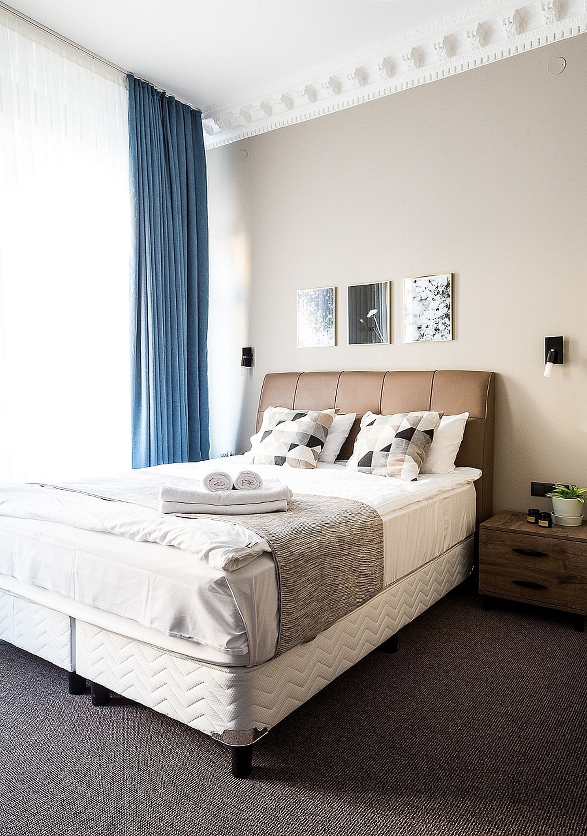 Una cama bien hecha con edredón blanco y almohadas, una mesita de noche con una planta, y una pared con tres cuadros enmarcados.