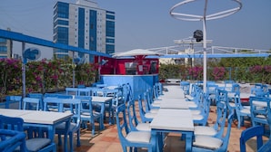 An outdoor seating area with blue tables and chairs, a fan, and a building in the background. - Evergreen Hotel (Mumbai)