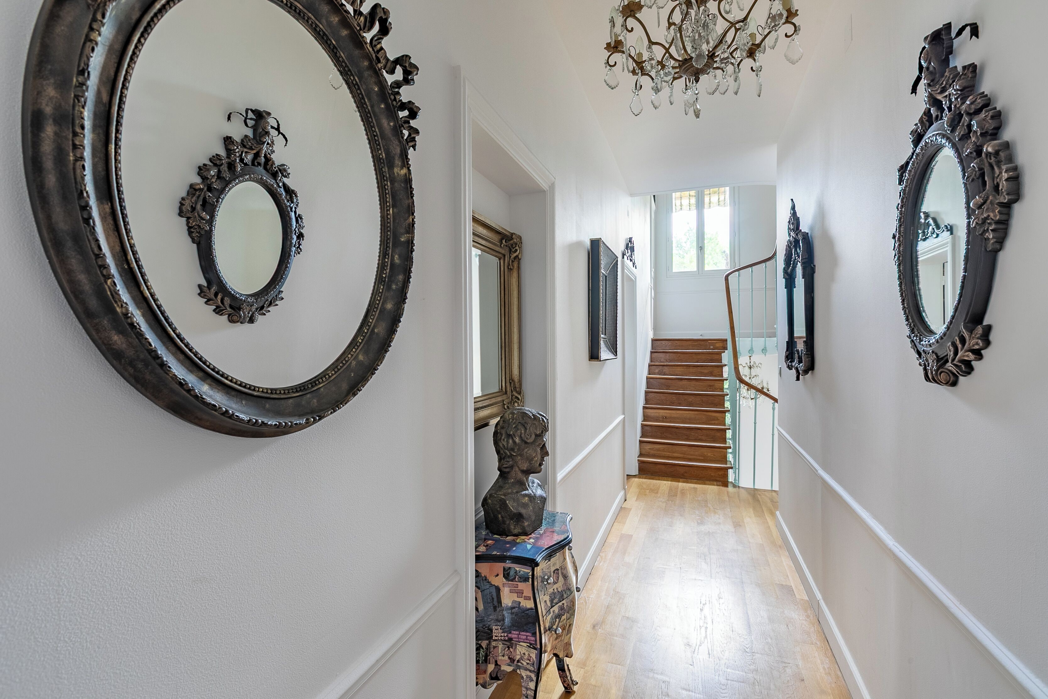 A hallway with ornate mirrors, a bust sculpture, and a chandelier.