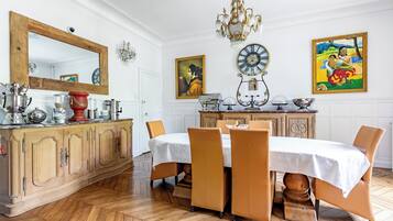 Un comedor con piso de madera en patrón de espiga, una mesa blanca cubierta con mantel blanco y sillas tapizadas en tono café.