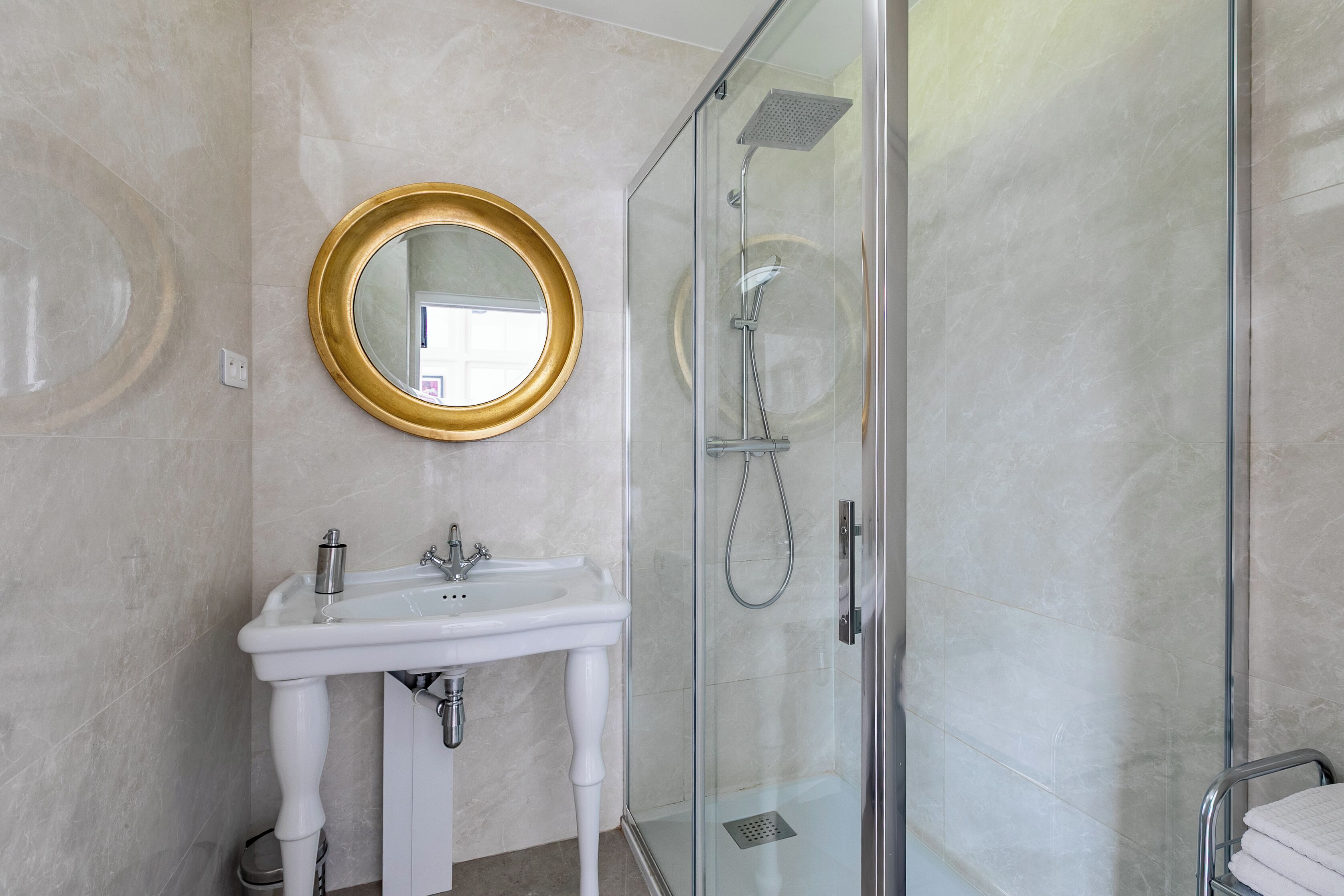 Een badkamer met een witte wastafel, een ronde spiegel met gouden lijst en een douche met glazen wanden.