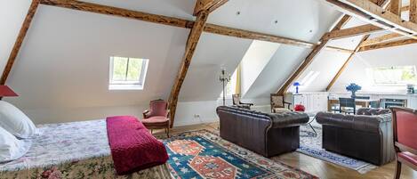 A loft bedroom with a sloped ceiling, wooden beams, a bed with floral bedding, a red throw blanket, a patterned rug, and a seating area with leather sofas and a chair.