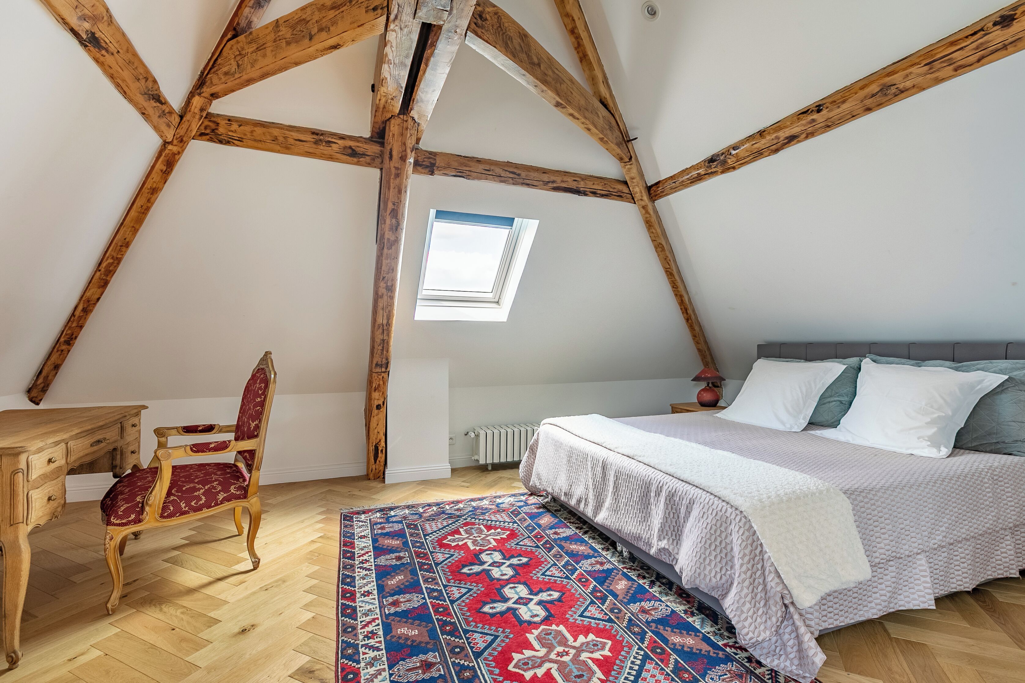 A bedroom with a sloped ceiling, a wooden desk, a red upholstered chair, a patterned rug, and a bed with white bedding.