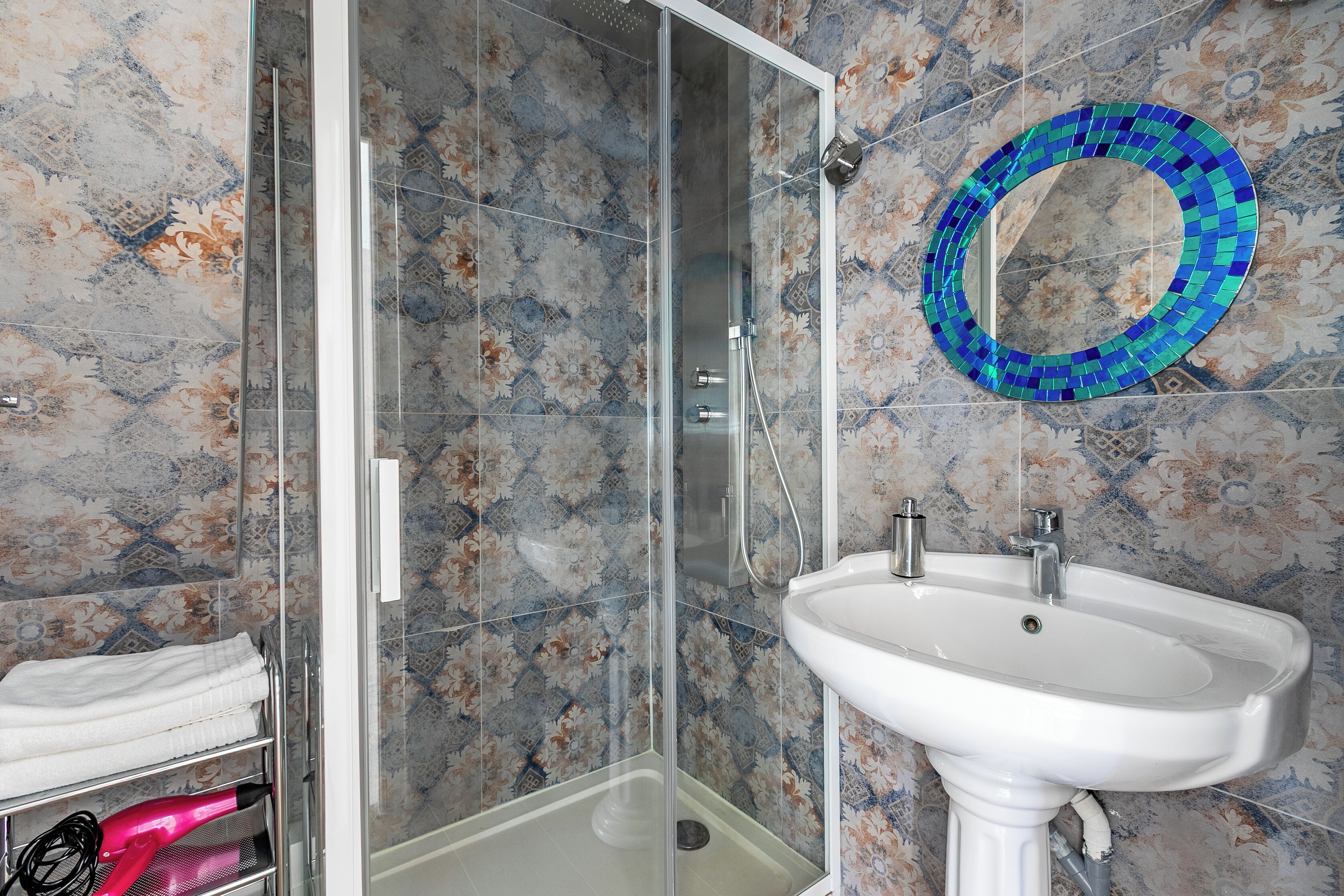 Un baño con cabina de ducha de vidrio, un lavamanos blanco y un espejo decorativo con mosaico azul.