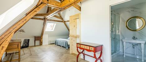 Una habitación con techo inclinado, vigas de madera, suelo de espiga, cama, escritorio, silla y baño con espejo y lavabo.